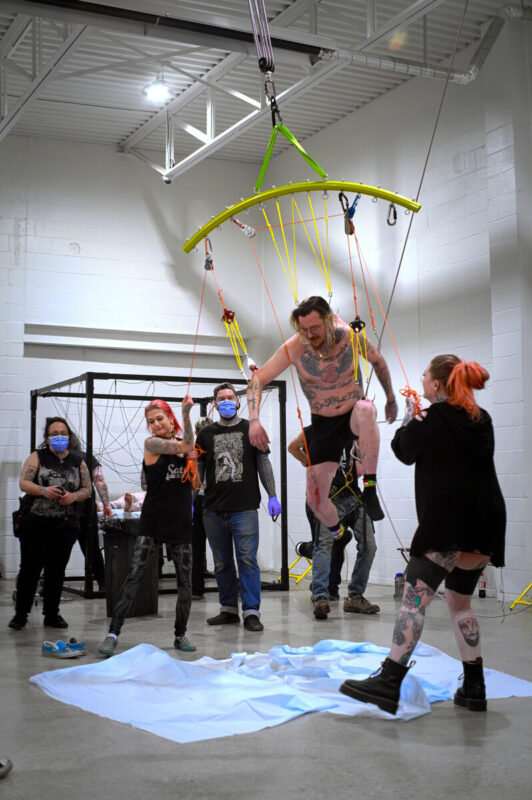 A person suspends off the ground, from several hooks on a large metal rig, with a group of people around them, watching.